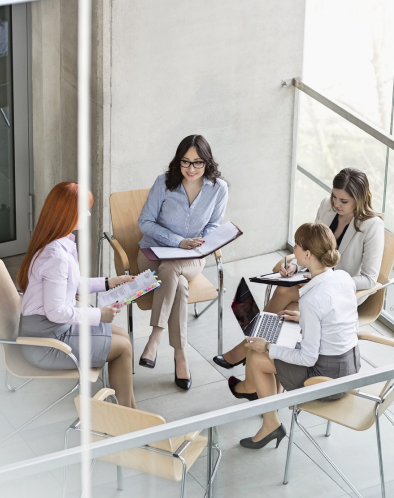 A group of five women in a bright, modern room, seated in a circle and talking, with notebooks and papers. No pink line